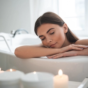 A woman relaxes in a bathtub, leaning her head on crossed arms, surrounded by lit candles in a peaceful bathroom setting.